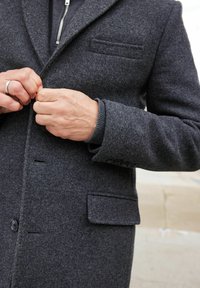 Abrigo de lana gris oscuro con cuello de muesca, cierre de botón en la parte delantera y un bolsillo en el pecho. Los puños cuentan con tejido de punto y acentos de botones.
