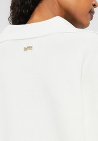 Sweater branco de malha com colarinho e um pequeno logótipo dourado "Barbour International" na parte de trás, com uma textura suave e um design simples.