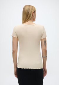 Γυναίκα με μακριά ξανθά μαλλιά  που φοράει μπεζ κοντομάνικη μπλούζα με ρίγες και μαύρη φούστα, εμφανίζοντας τα τατουάζ στα χέρια της, με την πλάτη στη λευκή φόντο.