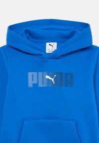 Modrá mikina s kapucňou a predným kengurím vreckom, s logom Puma zloženej z gradientných bodiek po celom hrudníku.