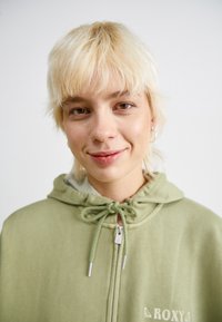 Zelená mikina na zips s kapucňou a šnúrkou na stiahnutie, s jemnou textúrou a svetlou farbou. Detail loga v dolnom rohu.
