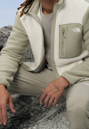 Homme portant une veste et un pantalon en polaire beige bicolore, assis sur un rocher gris, montrant un tatouage sur la main gauche.