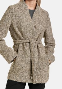 Beżowy płaszcz z teksturą i kołnierzem szalowym, z dwiema przednimi kieszeniami, paskiem w talii oraz zapięciem na jeden guzik. Kobieca sylwetka.