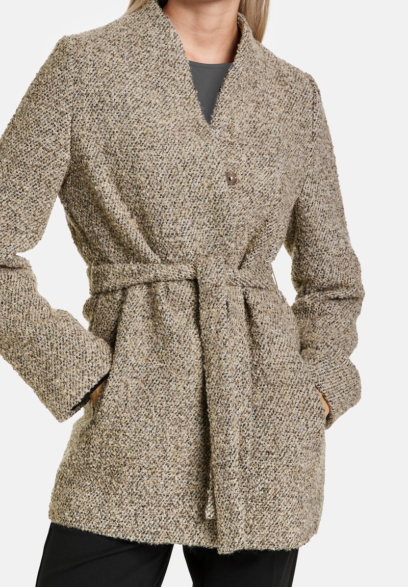Beżowy płaszcz z teksturą i kołnierzem szalowym, z dwiema przednimi kieszeniami, paskiem w talii oraz zapięciem na jeden guzik. Kobieca sylwetka.