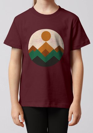 Kind draagt een bordeauxrode t-shirt met een cirkelvormig geometrisch ontwerp van bergen en zon in bruin, groen en zwart.