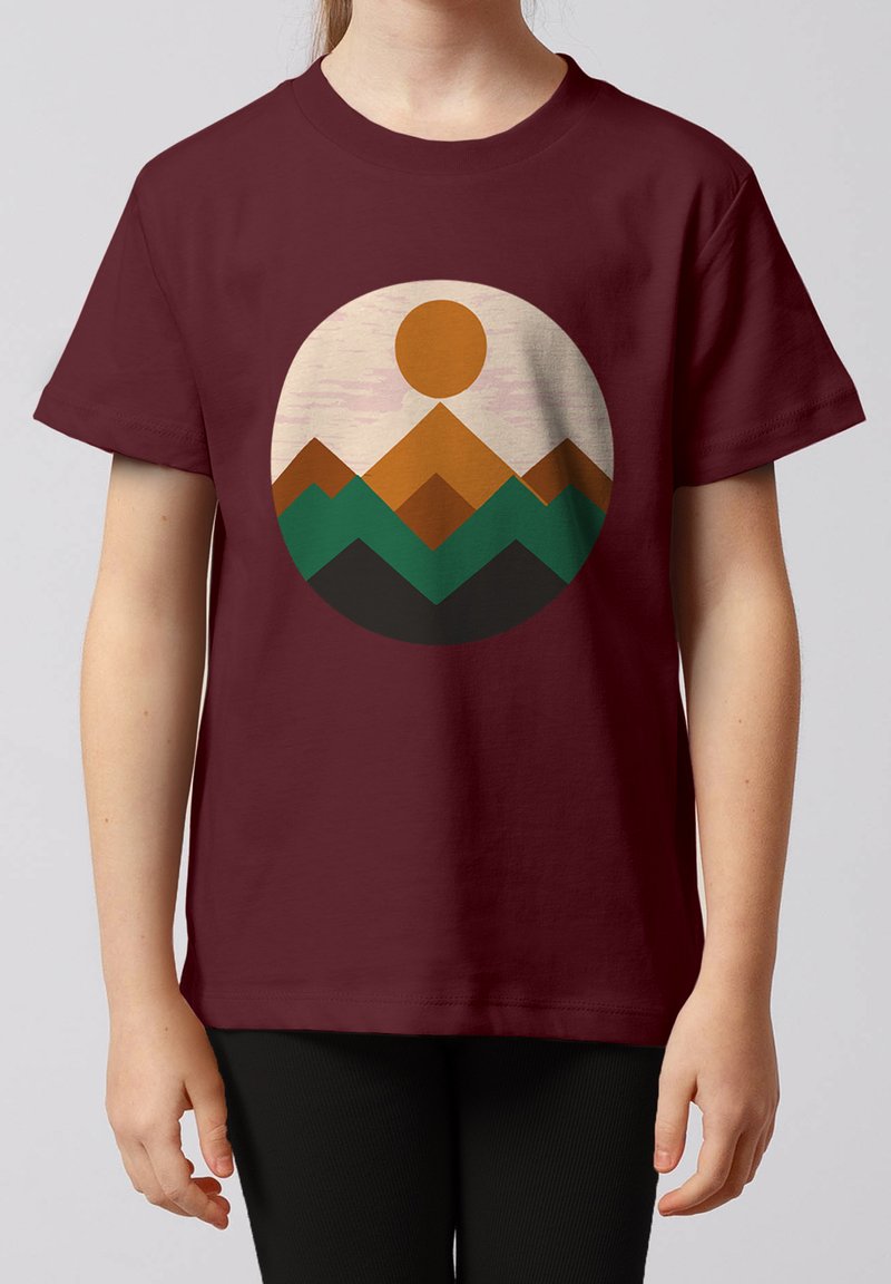 Enfant portant un t-shirt bordeaux avec un motif géométrique circulaire représentant une montagne et un soleil en couleurs marron, vert et noir.