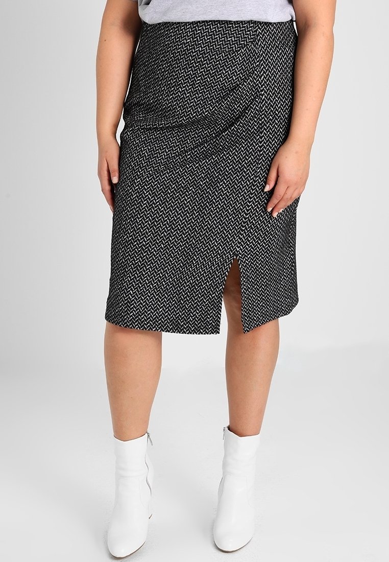 Femme portant une jupe noire et blanche à motifs, longueur genou avec une fente devant, et des bottines blanches, sur un fond uni.