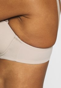 Beige bh med slät tyg, bred resår, justerbara band och en sömlös design. Visar närbild av sidostrukturen och sömmarna.