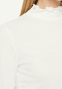 Vit långärmad topp med ribbad textur, har en volangkrage. Tyget ser mjukt ut med en lätt lyster.