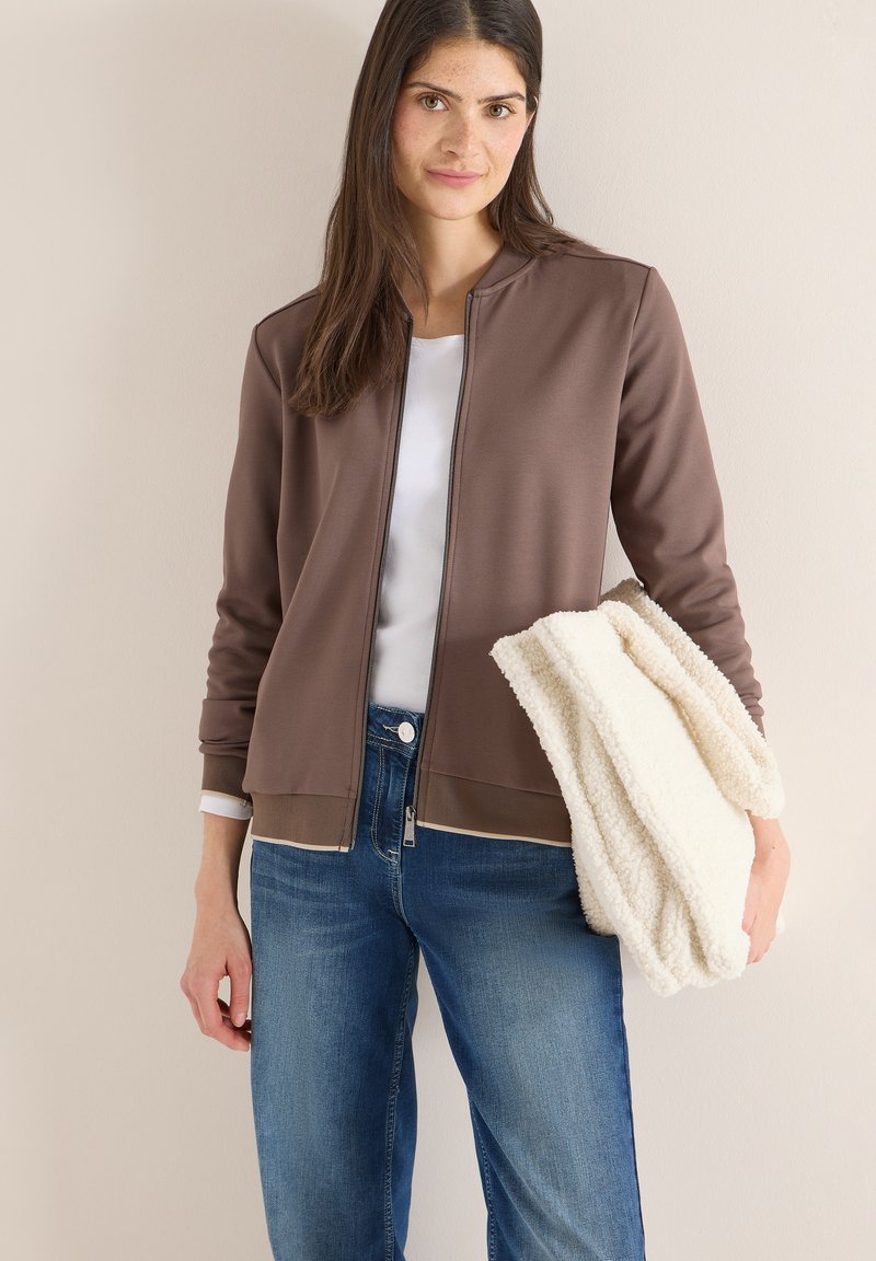 Femme portant une veste marron à fermeture éclair sur une chemise blanche et un jean bleu, tenant une couverture crème texturée pliée.