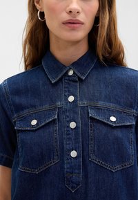 Temno modra denim srajca s kratkimi rokavi, zapenjanjem s sprednjimi gumbi, ostrim ovratnikom in dvema prsnikoma. Srebrni sponki in vidni šivi.