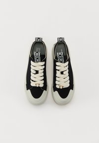 Sorte kanvas sneakers med hvite snørebånd, en gullakssent på tungen, og en teksturert lys grå såle. Har en mønstret løkkebånd.