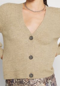 Cardigan de punto beige con escote en V, cinco botones grandes y textura acanalada. Presenta mangas cortas y un estilo corto.