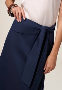 Marineblauwe wikkelrok met een brede tailleband en strikdetail. Soepele stof textuur, gecombineerd met een lichtgekleurde blouse. Armband om de pols.