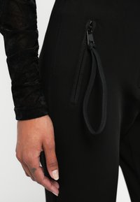 Črne športne hlače z zadrgo in tekstilnim trakom, v kombinaciji z teksturirano črno srajco z dolgimi rokavi. Poudarek na podrobnostih roke.