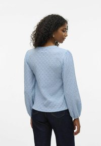 Blusa leggera di colore azzurro con texture, maniche lunghe a sbuffo, scollatura rotonda e una vestibilità rilassata, abbinata a jeans in denim scuro.