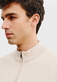Beżowa bluza rozpinana na zamek z wysokim, prążkowanym kołnierzem i teksturowanym materiałem, z metalowym zamkiem błyskawicznym i gładką powierzchnią.