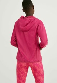 Personne portant une veste à capuche rose vif et des leggings à motif rose, debout les mains sur les hanches, dos tourné, sur un fond uni.