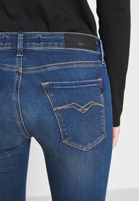 Jeans de mezclilla azul oscuro con un bolsillo trasero que presenta un diseño de costura en zigzag único y un parche de piel negra de la marca en la cintura.