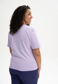 Helllila Baumwoll-T-Shirt mit kurzen Ärmeln, lockerer Passform und rundem Ausschnitt, von hinten präsentiert. Glatte Textur, ohne Muster.