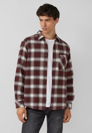 Jonge man draagt een open rood-wit geruit overhemd over een wit T-shirt en zwarte jeans, kijkt zijwaarts tegen een effen achtergrond.