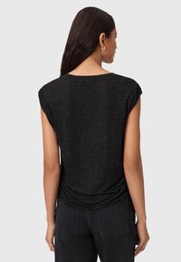 Donna con lunghi capelli scuri che indossa una maglietta nera a maniche corte con macchie e jeans scuri, vista di spalle su uno sfondo neutro.