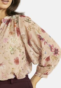 Ziedu blūze maigā rozā audumā ar vaļīgu piegriezumu, savāktām piedurknēm un zelta akcentiem, izceļot dažādas krāsainu ziedu rakstus.