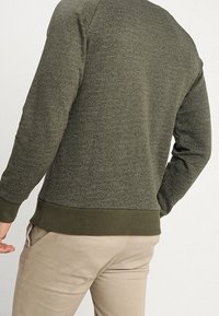 Felpa verde oliva con un motivo testurizzato, polsini e orlo a costine. Presenta un collo rotondo e una silhouette aderente.