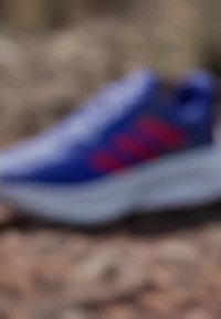 Blue athletic shoe with red accents, featuring a textured upper and thick white sole. Laces are visible, and the design is modern and streamlined.