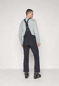 Pantalones de esquí negros con tirantes ajustables, bolsillos laterales y un ajuste relajado. Llevados sobre una camiseta de manga larga gris claro y botas de esquí.