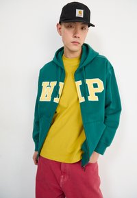 Sweat à capuche zippé vert avec des lettres "WIP" jaunes, associé à un t-shirt jaune et à un pantalon rose. Le modèle porte une casquette noire.