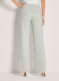 Vrouw in hoge taille, wijde lichtgrijze geblokte broek en witte hoge hakken, staande tegen een effen muur.