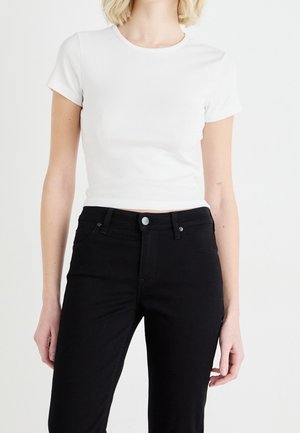 Vrouw draagt een aansluitend wit crop top met korte mouwen, gecombineerd met een zwarte, hooggetailleerde jeans, tegen een effen achtergrond.