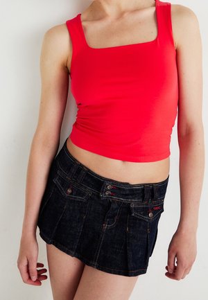 Kobieta w czerwonym, bezrękawnikowym topie i ciemnoniebieskiej mini spódniczce, stojąca na tle białej ściany.