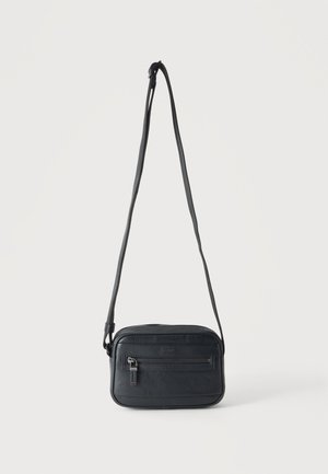 Zwarte crossbodytas met een voorvak met rits, een gestructureerd oppervlak en een verstelbare band, hangend tegen een effen lichte achtergrond.