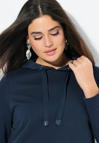 Felpa con cappuccio blu navy in tessuto testurizzato, con coulisse piatte e polsini a coste. Orecchini argento notevoli che aggiungono contrasto.