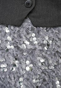 Pelēks tekstila augšdaļa ar vienu melnu pogu, uzlikta uz gaiši pelēkas, franču malām apstrādātas auduma ar apaļām sudraboti spīdošām fliterēm.