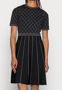 Robe en maille noire à manches courtes, avec un motif en losange et des coutures contrastantes. Silhouette trapèze avec des détails en panneaux verticaux.