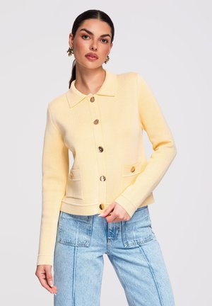 Femme portant un cardigan boutonné jaune pâle avec des poches avant et un jean taille haute bleu clair, sur un fond uni.