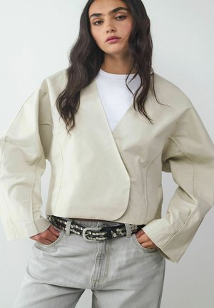 Hellbeige, taillierte Jacke mit Wickeldesign, weiten Ärmeln und glatter Textur, kombiniert mit hochgeschnittenen grauen Jeans und einem genadelten schwarzen Gürtel.