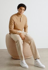 Camisa polo de manga larga beige, pantalones crema y zapatillas blancas. Sentado en un ottoman redondo y texturizado beige contra un fondo liso.