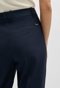 Pantaloni blu navy su misura realizzati con un tessuto testurizzato, caratterizzati da una singola tasca posteriore e un accento con logo dorato. Vestibilità classica con passanti per cintura.