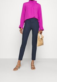 Žena v jasně fialové blůze s dlouhým rukávem a úzkých navy kalhotách, držící béžovou kabelku a stojící na béžových sandálech na vysokém podpatku.