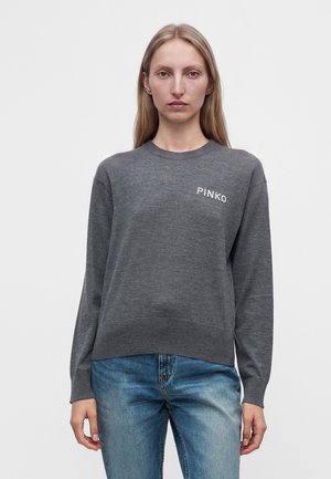 Frau mit langen blonden Haaren, die einen grauen Pullover mit dem Text "PINKO" und blaue Jeans trägt, steht vor einem einfarbigen hellen Hintergrund.