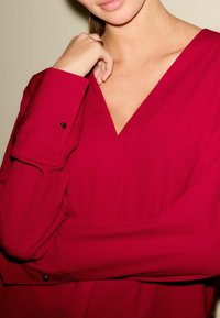 Raudona bluzonė su V formos iškirpte, ilgomis rankovėmis ir užsegamais rankogaliais. Pagaminta iš lygaus audinio su švelniu blizgesiu. Yra paslėptas kišenė.