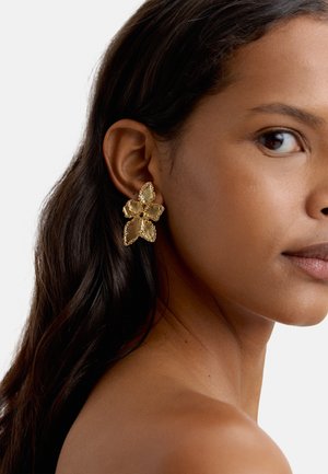 Zbliżenie kobiety noszącej duże złote kolczyki w kształcie kwiatu z teksturowanymi płatkami, ukazujące prawy bok jej twarzy i ramienia.