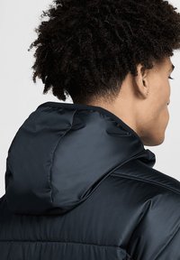 Marineblaue Pufferjacke mit einer weichen, glänzenden Textur, ausgestattet mit Kapuze und gestepptem Design, von hinten präsentiert, wobei strukturiertes Haar sichtbar ist.