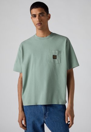 Junger Mann trägt ein hellgrünes kurzärmeliges T-Shirt mit Brusttasche und blaue Jeans vor einem einfarbigen hellgrauen Hintergrund.