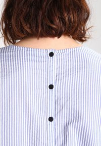 Blusa a righe blu e bianche con scollatura rotonda e bottoni neri in plastica lungo la schiena. Tessuto leggero con una texture sottile.