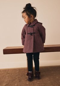 Manteau à motif pied-de-poule rouge et noir avec des fermetures à bascule noires, porté par-dessus des leggings noirs et des chaussures bordeaux brillantes avec des accents de nœuds.